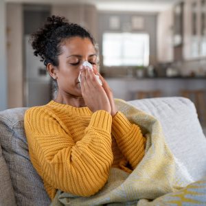 Portrait of young black woman sneezing in to tissue at home. Sick african woman wrapped in blanket sitting on sofa blowing her nose at home. Ill girl sneezing with runny nose in winter.